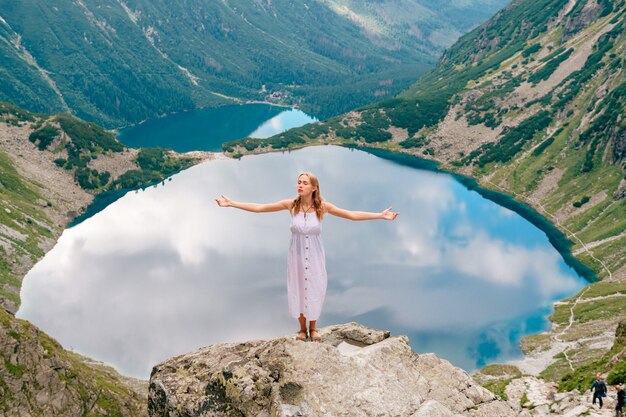 Joven rubia en vestido blanco de pie en piedra en las montañas con las manos separadas y hermoso lago detrás.