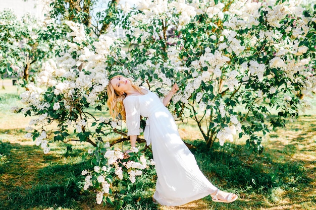 Joven rubia con vestido blanco posando en arbustos de lilas