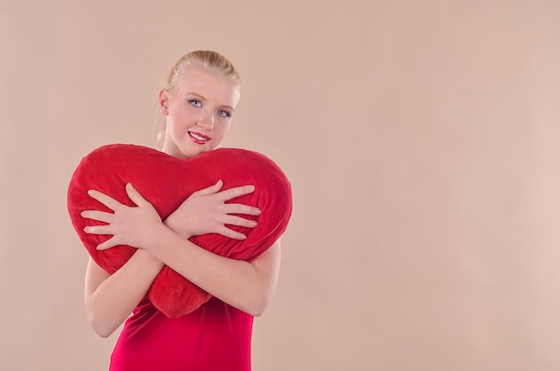 Una joven rubia con un vestido rojo sostiene un corazón rojo en sus manos con un fondo beige