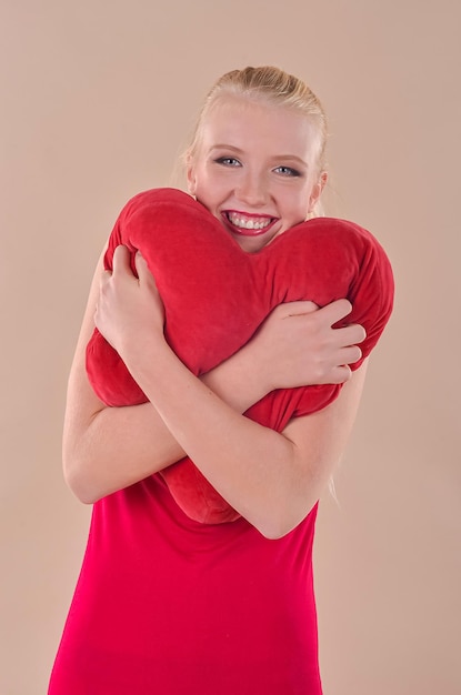 Una joven rubia con un vestido rojo sostiene un corazón rojo en sus manos con un fondo beige