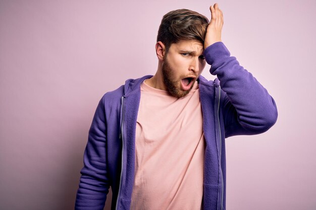 Joven rubio con barba y ojos azules con sudadera morada sobre fondo rosa sorprendido con la mano en la cabeza por error recuerda error Olvidó el concepto de mala memoria