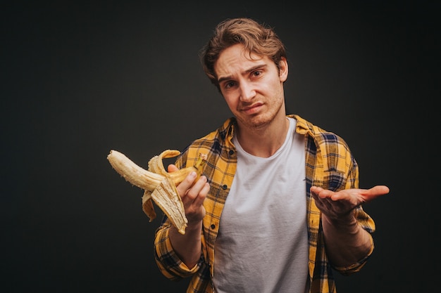 Joven rubio con camisa amarilla está comiendo plátano dudosamente