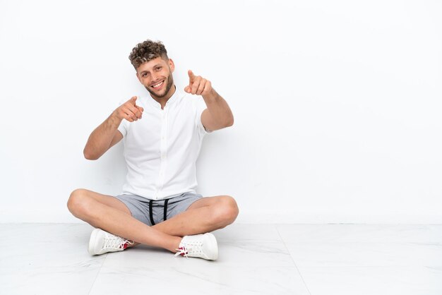 Foto un joven rubio sentado en el suelo aislado de fondo blanco te señala con el dedo mientras sonríe