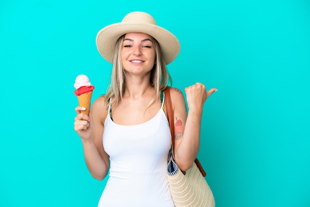 Joven rumana sosteniendo helado y bolsa de playa aislada de fondo azul apuntando hacia un lado para presentar un producto