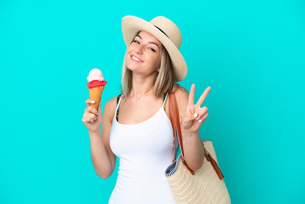 Joven rumana sosteniendo helado y bolsa de playa aislada de fondo azul sonriendo y mostrando el signo de la victoria