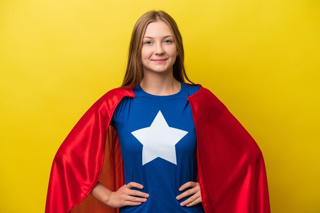 Foto joven rusa aislada de fondo amarillo con traje de superhéroe posando con los brazos en la cadera y sonriendo