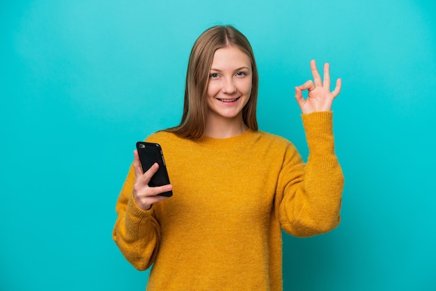 Joven rusa aislada de fondo azul usando teléfono móvil y haciendo el signo de OK