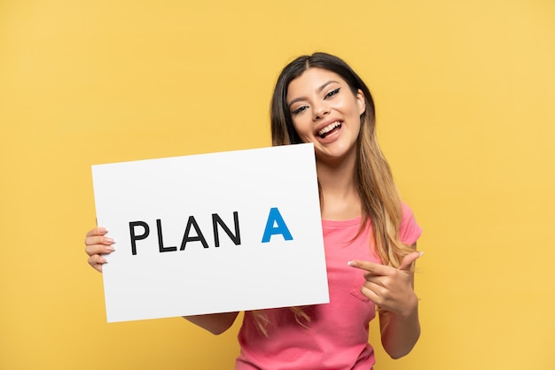 Joven rusa aislada sobre fondo amarillo sosteniendo un cartel con el mensaje PLAN A y apuntando