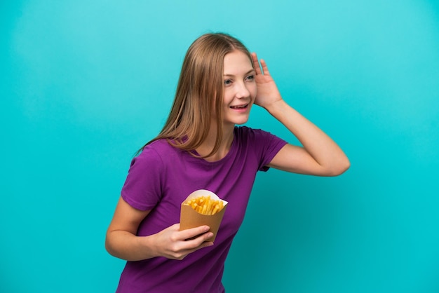 Joven rusa atrapando papas fritas aisladas de fondo azul escuchando algo poniendo la mano en la oreja