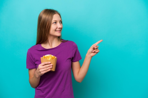Joven rusa atrapando papas fritas aisladas de fondo azul señalando con el dedo al costado y presentando un producto