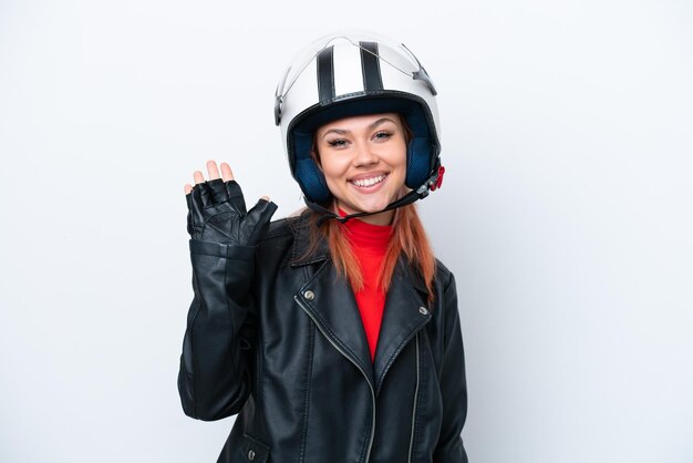 Joven rusa con un casco de motocicleta aislado de fondo blanco saludando con la mano con expresión feliz