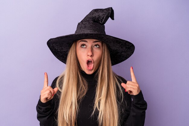 Joven rusa disfrazada de bruja celebrando Halloween aislado sobre fondo púrpura apuntando al revés con la boca abierta.