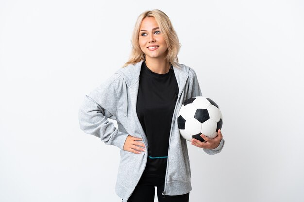 Joven rusa jugando al fútbol aislado en la pared blanca posando con los brazos en la cadera y sonriendo