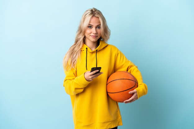 Joven rusa jugando baloncesto aislado en azul enviando un mensaje con el móvil