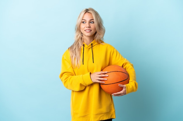 Joven rusa jugando baloncesto aislado en la pared azul mirando hacia el lado y sonriendo