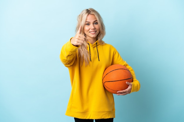 Joven rusa jugando baloncesto aislado en la pared azul con los pulgares hacia arriba porque ha sucedido algo bueno