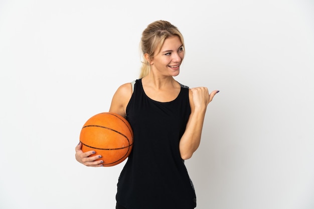 Joven rusa jugando baloncesto aislado en la pared blanca apuntando hacia el lado para presentar un producto