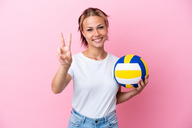 Joven rusa jugando voleibol aislada de fondo rosa sonriendo y mostrando el signo de la victoria