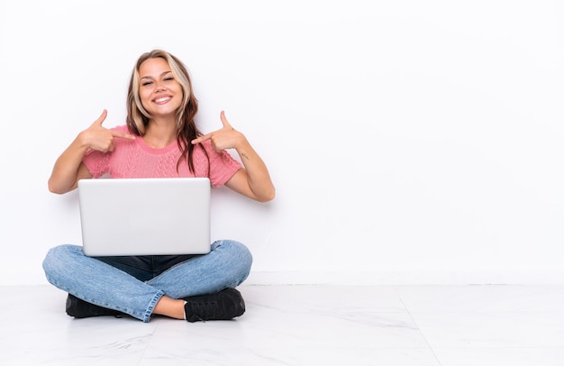 Joven rusa con una laptop sentada en el suelo aislada de fondo blanco orgullosa y satisfecha