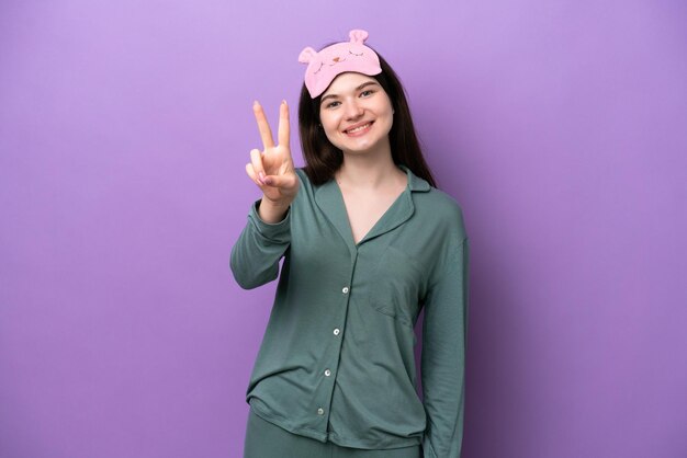 Joven rusa en pijama aislada de fondo morado sonriendo y mostrando el signo de la victoria