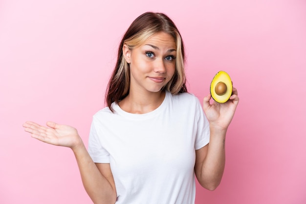 Joven rusa sosteniendo aguacate aislado de fondo rosa con dudas mientras levanta las manos