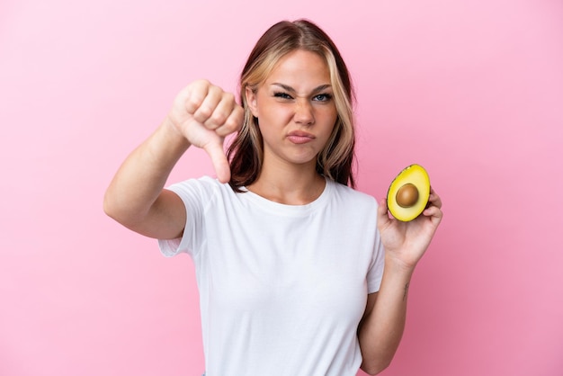 Joven rusa sosteniendo aguacate aislado de fondo rosa mostrando el pulgar hacia abajo con expresión negativa