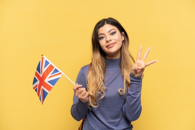 Joven rusa sosteniendo una bandera del Reino Unido aislada sobre fondo amarillo feliz y contando tres con los dedos