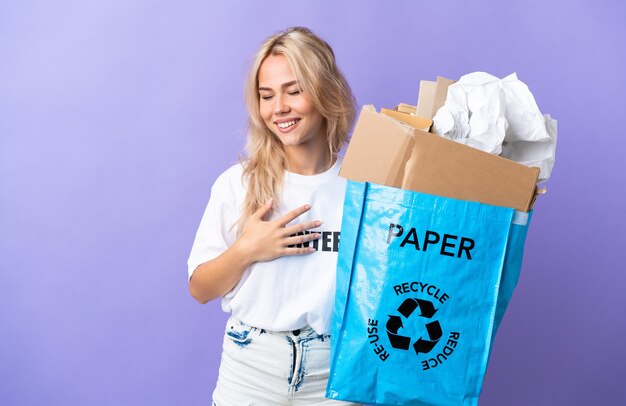 Joven rusa sosteniendo una bolsa de reciclaje llena de papel para reciclar aislado en púrpura sonriendo mucho