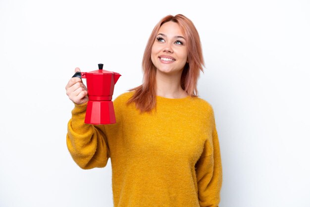 Joven rusa sosteniendo una cafetera aislada de fondo blanco mirando hacia arriba mientras sonríe