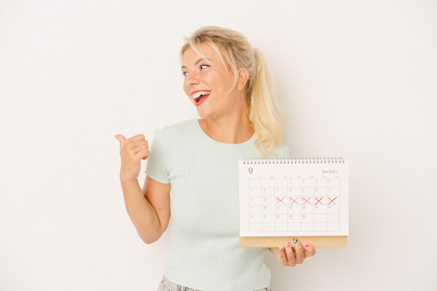 Joven rusa sosteniendo un calendario aislado sobre fondo blanco apunta con el dedo pulgar lejos, riendo y despreocupado.