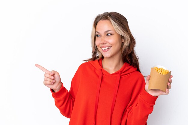 Joven rusa sosteniendo patatas fritas aisladas de fondo blanco señalando con el dedo a un lado y presentando un producto