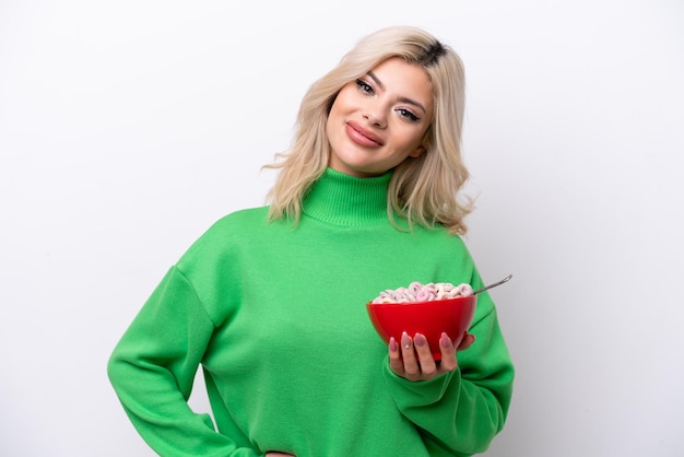 Joven rusa sosteniendo un plato de cereales aislado de fondo blanco posando con los brazos en la cadera y sonriendo