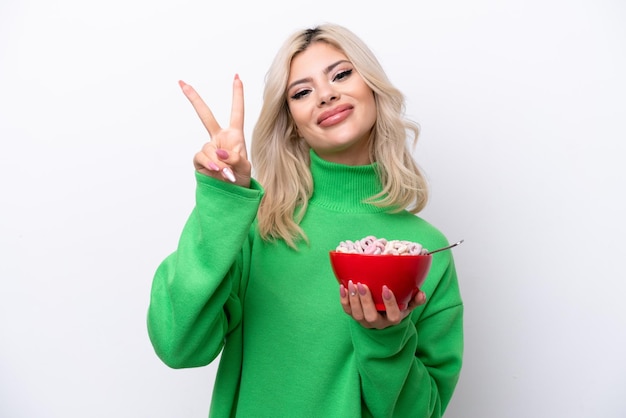 Joven rusa sosteniendo un plato de cereales aislado de fondo blanco sonriendo y mostrando el signo de la victoria