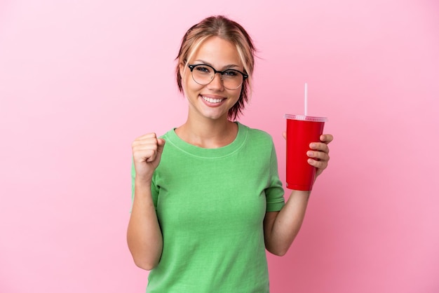 Joven rusa sosteniendo un refresco aislado de fondo rosa celebrando una victoria en posición ganadora