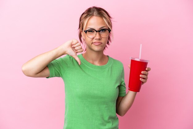 Foto joven rusa sosteniendo un refresco aislado de fondo rosa mostrando el pulgar hacia abajo con expresión negativa