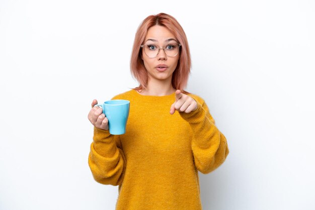 Joven rusa sosteniendo una taza de café aislada de fondo blanco sorprendida y apuntando al frente