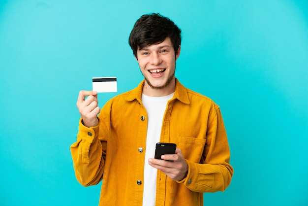 Joven ruso aislado sobre fondo azul comprando con el móvil con tarjeta de crédito