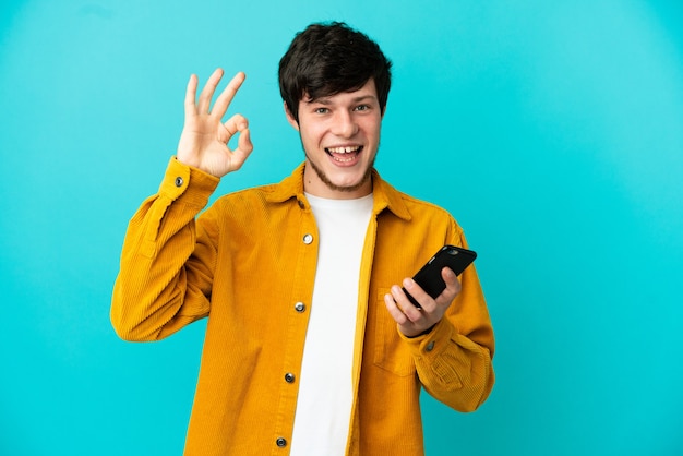 Joven ruso aislado sobre fondo azul con teléfono móvil y haciendo el signo de OK