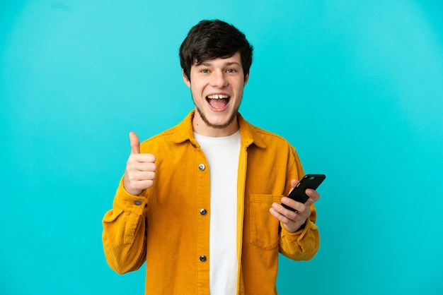 Joven ruso aislado sobre fondo azul con teléfono móvil mientras hace los pulgares para arriba