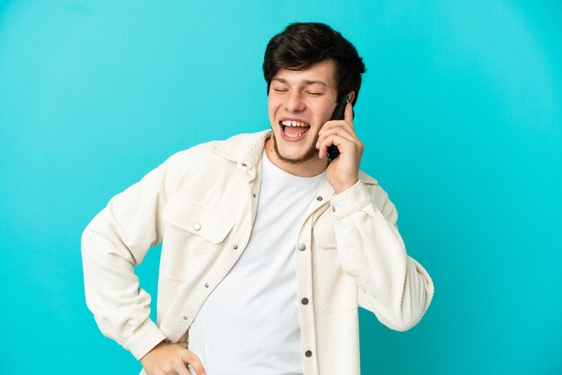 Joven ruso con teléfono móvil aislado sobre fondo azul posando con los brazos en la cadera y sonriendo