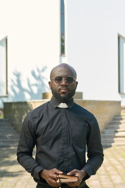 Joven sacerdote serio con camisa negra con cuello clerical sosteniendo la sagrada biblia