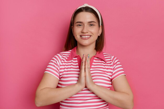 Una joven satisfecha y guapa con una camiseta informal a rayas suplica piedad tiene una expresión positiva una sonrisa suave mantiene las palmas en gesto de oración pide un deseo de pie aislado en un fondo rosa