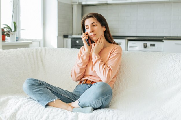 Foto una joven satisfecha hablando en su teléfono inteligente mientras está sentada en un acogedor sofá en su sala de estar la auténtica escena doméstica se completa con la cálida decoración