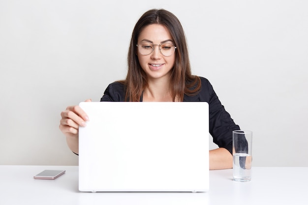 Una joven satisfecha se sienta frente a una computadora portátil abierta, mira un seminario web, piensa en crear un nuevo diseño web, usa gafas redondas para una buena visión, bebe agua, se sienta sola en interiores.