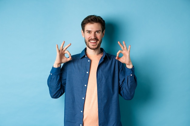 Joven satisfecho sonriendo y mostrando un gesto bien, aprueba algo bueno, alabando algo excelente, de pie sobre fondo azul.