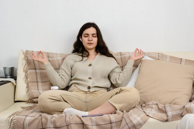 Una joven sentada en el sofá escucha la meditación con los ojos cerrados a través de los auriculares.