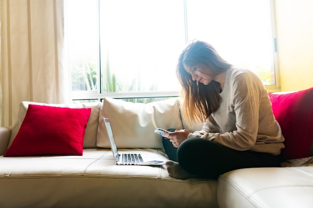 una joven sentada en el sofá frente a una gran ventana usando un teléfono inteligente y una computadora portátil