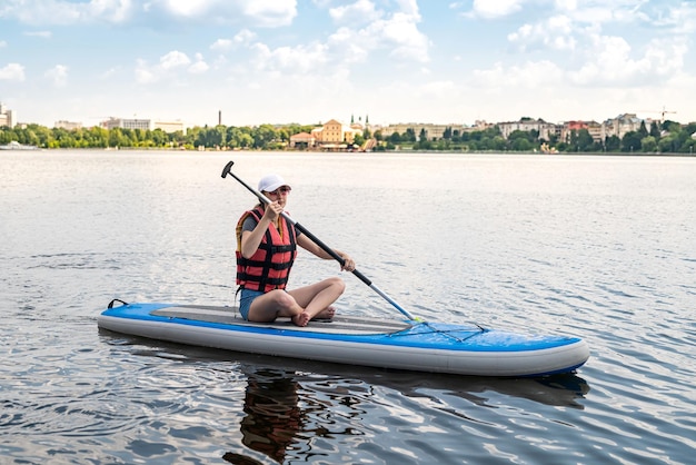 Una joven sentada en una tabla de remo usa un chaleco salvavidas y aprende a nadar un estilo de vida activo de verano