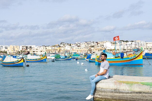 Joven sentado y al fondo el mar lleno de coloridos barcos en Marsaxlokk