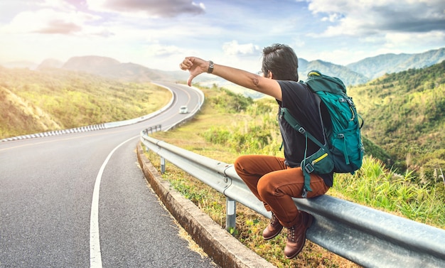 Joven sentado en la carretera. Conceptos de viaje y vacaciones. Mochilero en carretera. Hombre de viaje haciendo autostop